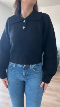 Pull en maille noir avec un col, trois boutons décoratifs et un ourlet côtelé. Porté avec un jean bleu clair, montrant une coupe décontractée.