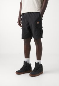 Black cargo shorts with side pockets and a logo patch, paired with high-top black sneakers and white crew socks on a neutral background.
