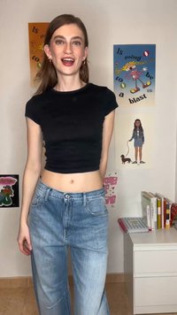 Jeune femme portant un t-shirt court noir et un jean bleu, souriante, debout dans une pièce avec des affiches colorées et une commode blanche avec des livres.