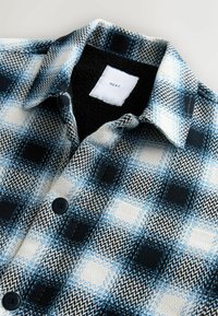 Košeľa z flanelu s károvaným vzorom v čiernej, modrej a bielej farbe. Má čiernu fleecovú podšívku a čierne gombíky na prednej strane.