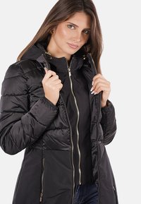 Giacca a vento nera con cappuccio, chiusura con zip frontale e tasche laterali. Caratterizzata da una texture trapuntata e dettagli in metallo dorato per chiusura e decorazioni.