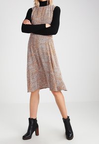 Hame ilman hihoja ruskeasta ja valkoisesta kuviollisesta kankaasta, A-linjan muotoilu, yhdistetty musta pitkähihainen paita ja tukevat mustat korkokengät.