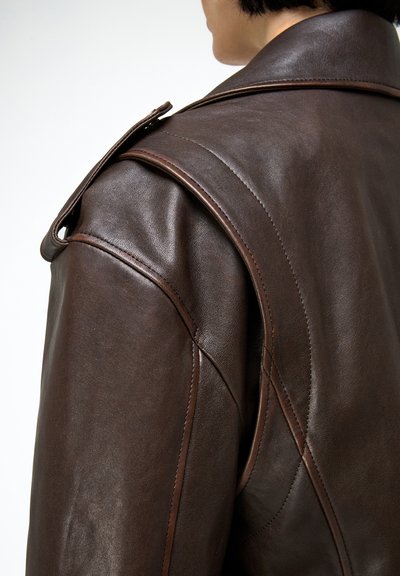 Sötétbarna bőrdzsekit viselő személy, részletes varrással és vállpánttal, egyszínű világos háttéren.