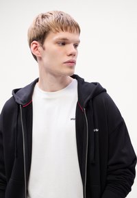 Čierna mikina s kapucňou na zips a červeným akcentom na kapucni, preložená cez biele tričko s nápisom "HUGO" vytlačeným čiernou farbou. Hladká textúra a moderný dizajn.