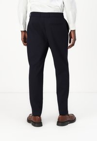 Pantaloni da abbigliamento navy con taglio dritto, vita slim, tasca posteriore e indossati con scarpe marroni; il tessuto appare liscio e su misura.