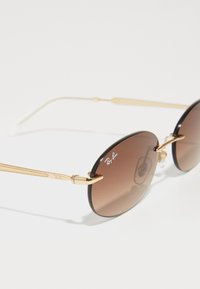 Auksinėmis rėmeliais Ray-Ban saulės akinius su apvaliais rudi gradientiniai objektyvais ir plonais šonais ant balto fono.