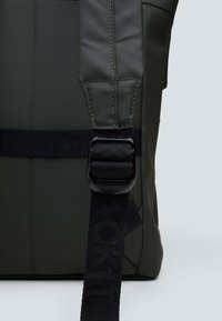 Vihreä reppu on valmistettu teksturoidusta, vesitiivistä materiaalista, ja siinä on leveä, säädettävä hihna sekä kestävä musta solki.