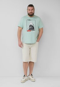 Mintgroene T-shirt met een haai grafiek, crèmekleurige shorts en beige sneakers met donkere accenten. Eenvoudig ontwerp, lichtgewicht katoen.