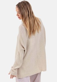 Pull en maille oversize beige clair avec manches côtelées et coupe décontractée. Texture douce et détails de coutures à l'arrière.