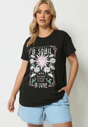Lyshåret kvinde iført sort T-shirt med sol, måne, tusindfryd og teksten "Le Soleil La Lune" samt lyseblå denimshorts.