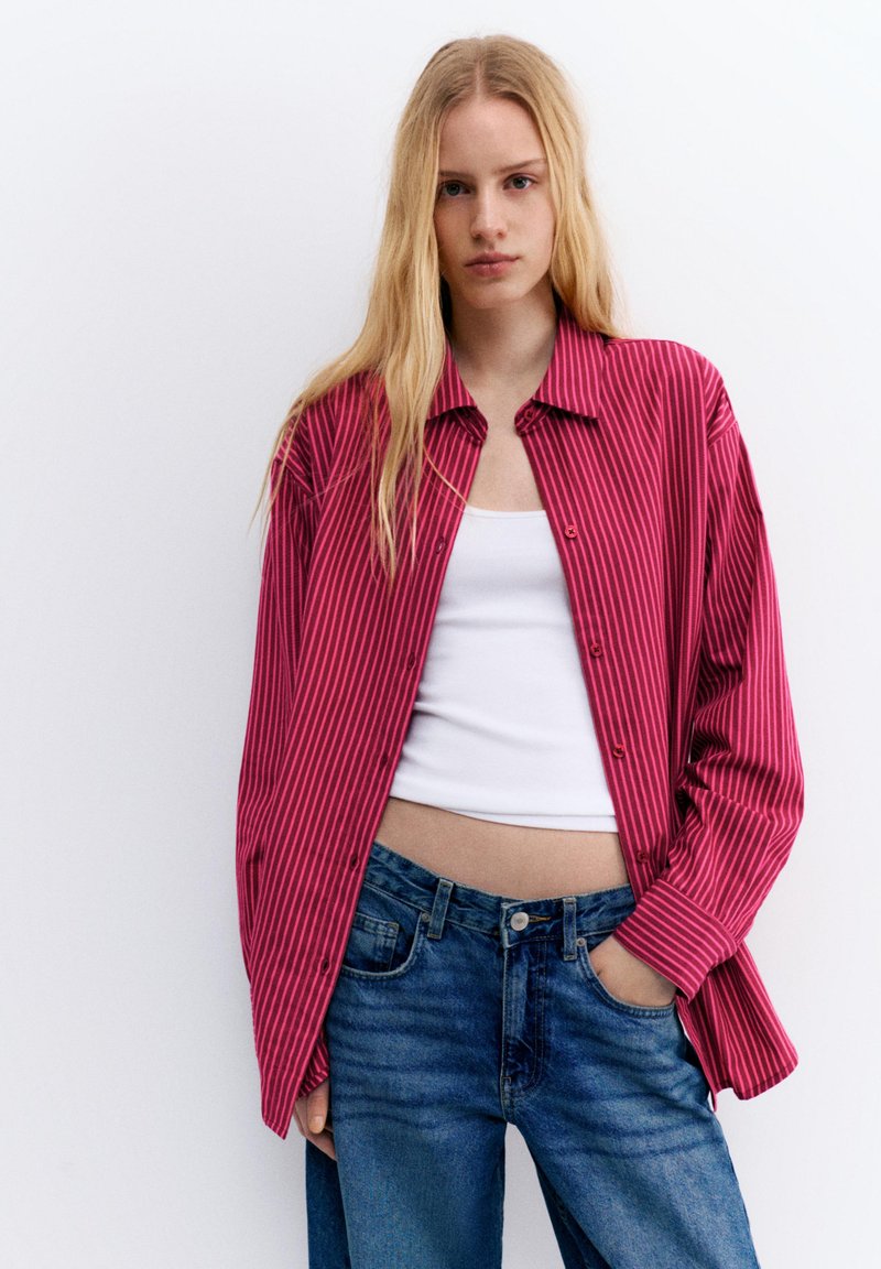 Ung kvinde med langt blondt hår iført en stor rød- og pinkstribet skjorte over en hvid crop top og blå jeans, stående med den ene hånd i lommen.