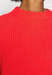 Rode gebreide trui met een gestructureerd ontwerp, een geribbelde halslijn en raglanmouwen, die een levendige kleur en gelijkmatige steekpatroon laat zien.