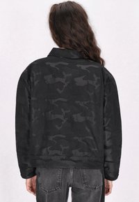 Zwarte camouflagejas met een relaxte pasvorm, voorzien van een kraag en lange mouwen. Glad textuur en subtiele glans.