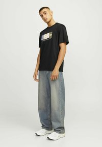 Camiseta de algodón negra con diseño gráfico frontal en varios colores, combinada con jeans de mezclilla azul claro sueltos y zapatillas blancas.