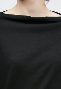 Μαύρο τοπ με λεία, ματ υφή, που διαθέτει φαρδύ, απαλά χαλαρωμένο ντεκολτέ. Ο σχεδιασμός τονίζει την απλότητα και τη μοντερνιά.