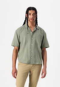 Camicetta a maniche corte, verde oliva, con bottoni e tasca sul petto, realizzata in tessuto testurizzato, con una vestibilità rilassata e un design del colletto.