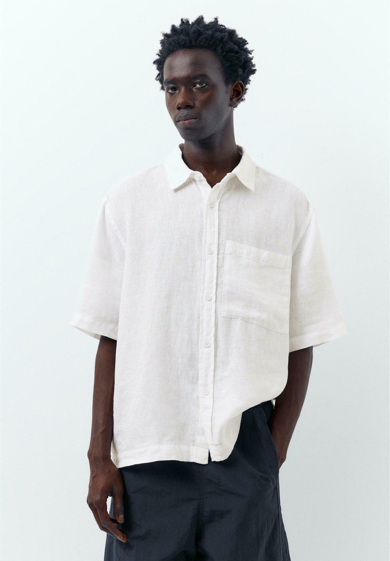 Jeune homme aux cheveux courts et bouclés portant une chemise blanche ample à manches courtes et un pantalon sombre, debout devant un fond blanc uni.