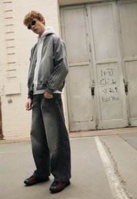 Grå bomberjakke med vertikale striper på ermene, hvit hettegenser under, oversized grå jeans og svart-røde sneakers. Urban bakgrunn.