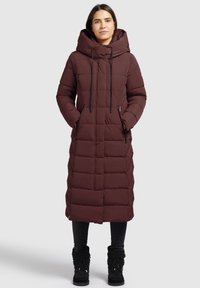 Burgunderlång pufferjacka med stor huva, dragkedjefickor, vertikalt quiltat mönster och snördetaljer, bärs över svarta leggings och stövlar.