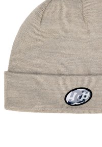 Czapka typu beanie wykonana z jasnobeżowej dzianiny o teksturowanym wykończeniu, z czarną owalną naszywką z białymi detalami z przodu.