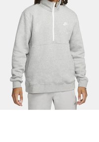 Muž v světlém šedém svršku Nike s čtvrtinovým zipem a odpovídajících světlých šedých kalhotách s logem Nike na stehně.