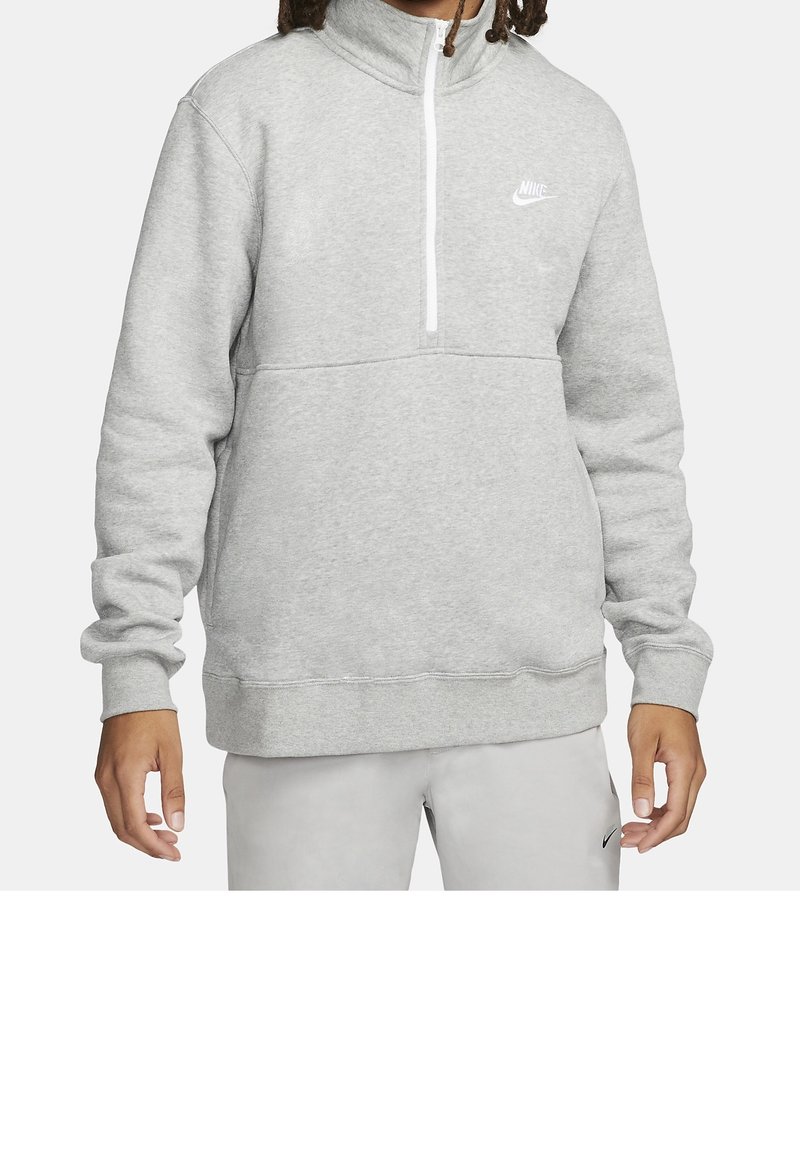 Muž v světlém šedém svršku Nike s čtvrtinovým zipem a odpovídajících světlých šedých kalhotách s logem Nike na stehně.