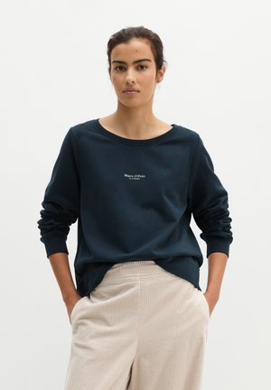 Femme aux cheveux foncés portant un sweat-shirt bleu marine avec un petit texte et un pantalon en velours côtelé beige clair, debout les mains dans les poches.