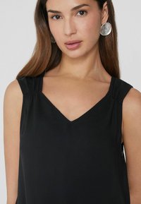 Zwarte mouwloze top met een V-halsontwerp, geplooide schouderdetails en een gladde textuur. Model draagt grote zilveren ronde oorbellen.
