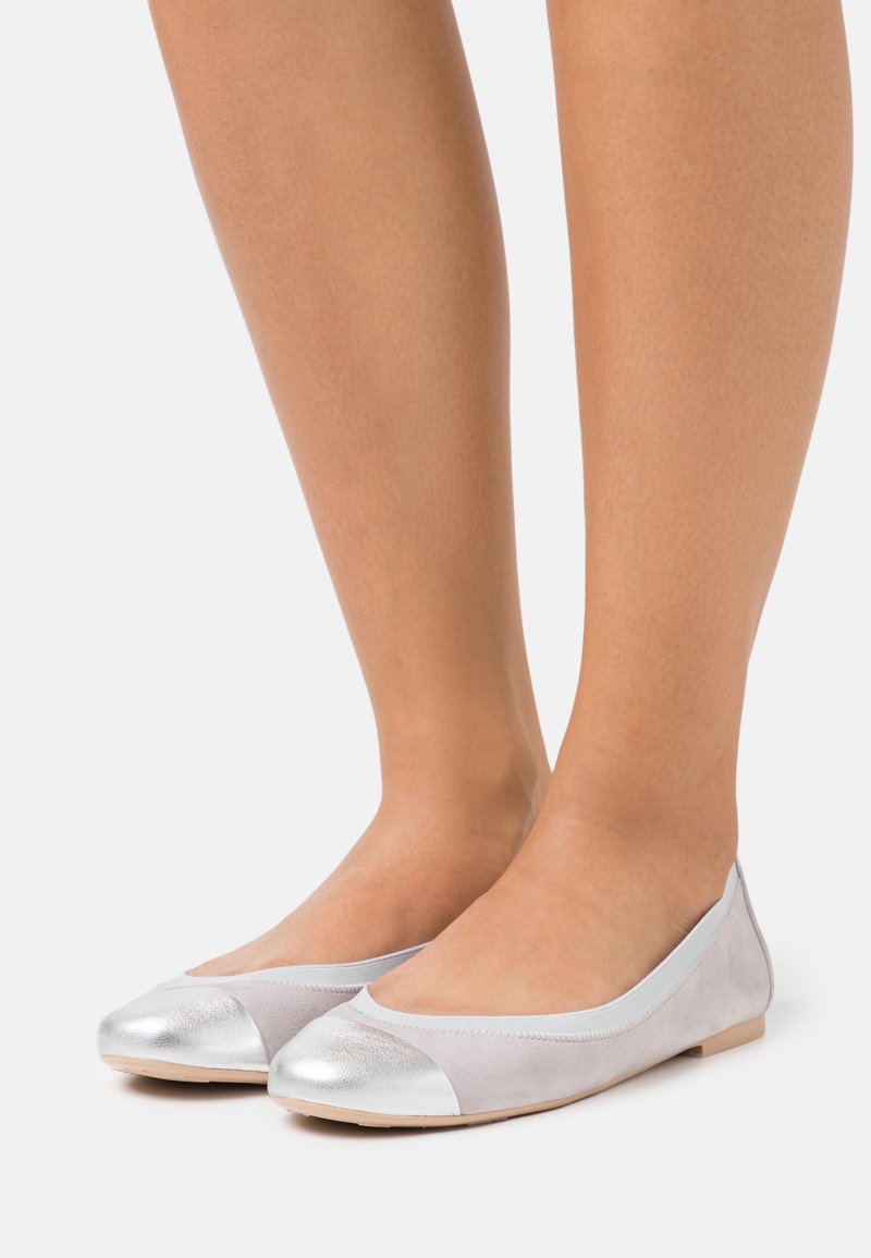 Jambes féminines portant des ballerines gris clair avec embouts métalliques argentés sur fond blanc.