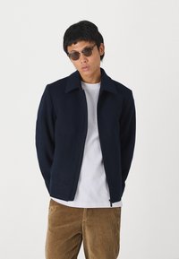 Giacca blu navy con zip e colletto, realizzata in un materiale simile alla lana. Indossata sopra una t-shirt bianca, abbinata a pantaloni in corduroy color sabbia.