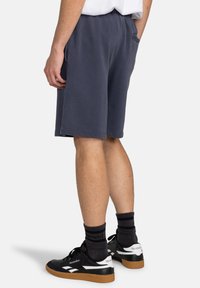 Gray cotton shorts with elastic waistband, side pockets, and a loose fit. Black sneakers with white accents and brown sole. Dark socks visible.