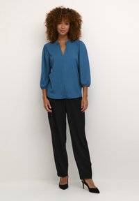 Blouse texturée teal avec un petit col, des manches bouffantes mi-longues et une fente sur le devant. Associée à un pantalon noir ample et des talons.