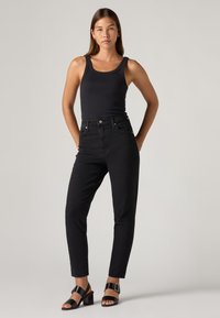 Mujer de pie con las manos en los bolsillos, vestida con una camiseta sin mangas negra, jeans negros de cintura alta y sandalias de tacón negras, ante un fondo liso.