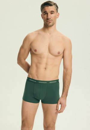 Mężczyzna bez koszulki o krótkich brązowych włosach, ubrany w ciemnozielone bokserki z paskiem na talii z napisem "Henderson", stojący na jednolitym jasnym tle.