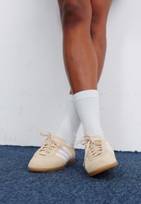 Svetlo bež semiš tenisice s tremi belimi črtami, ravnim svetlo rjavim podplatom in tankimi bež vezalkami, v kombinaciji z belimi rebrastimi nogavicami. 