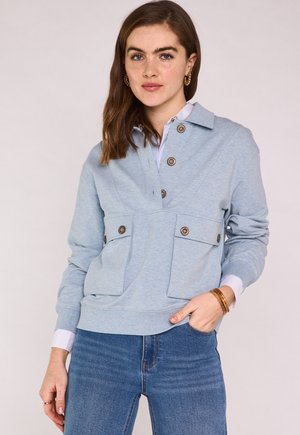 Donna con felpa azzurro chiaro bottonata con tasche a patta, jeans blu, orecchini a cerchio dorati e braccialetti, in piedi con una mano in tasca.