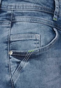 Jeans in denim azzurro chiaro con una texture scolorita, caratterizzati da molteplici tasche e cuciture a contrasto. Include un piccolo accento verde vicino alla tasca.