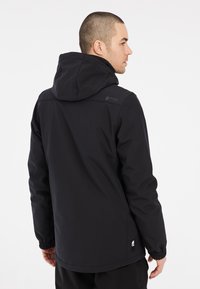 Veste noire imperméable avec capuche, poignets élastiques et ourlet ajustable. Présente un logo sur l'épaule et une texture lisse.