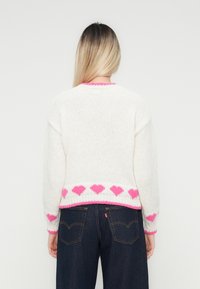 Sieviete ar gariem blondiem matiem, tērpusies baltā džemperī ar rozā sirdīm malās un uz aprocēm, ko papildina tumši zilas denima bikses, apgriezusies pretējo pusi.