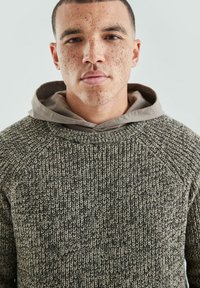 Sweter z dzianiny w kolorach ziemi, z teksturowanym wzorem, okrągłym dekoltem i wielowarstwowym beżowym kapturem. Miękki, swobodny krój z raglanowymi rękawami.