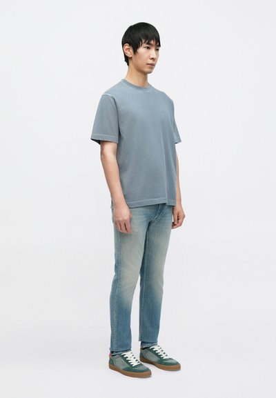 Ανοιχτό μπλε βαμβακερό T-shirt με κοντά μανίκια, συνδυασμένο με ξεθωριασμένο μπλε τζιν και πράσινα αθλητικά παπούτσια με λευκά κορδόνια και καφέ σόλες.