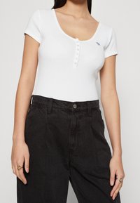 Hvid ribbet bodysuit med dyb udskæring og korte ærmer, der har en række af fem knapper, parret med højtaljede sorte bukser.