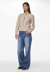 Beige cardigan, strikket tekstur, rund halslinning, frontknapper. Kombinert med vide blå jeans og svarte sko. Enkel, uformell design.
