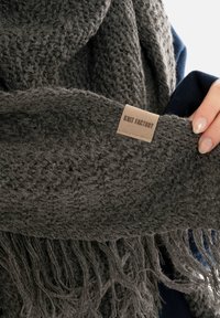 Donkergrijze gebreide sjaal met franjes aan de randen, voorzien van een textuurpatroon. Een beige label met de tekst "Knit Factory" is bevestigd.