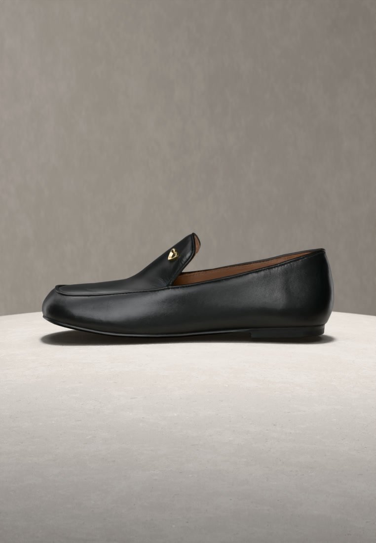 Mocassin en cuir noir à enfiler avec un petit détail cœur doré sur le côté, présenté sur une surface claire avec un fond neutre.