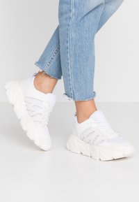 White athletic shoes featuring a mesh upper, prominent rubber sole, and wave-like design. The laces are threaded through looped accents.