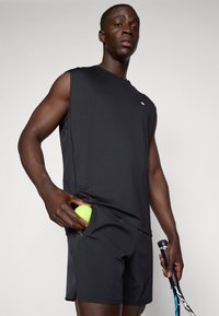 Čierny športový bezrukávový top a šortky, so bočnou kapsou, v ktorej sa nachádza tenisová loptička. Dizajn má hladkú textúru a decentné logo.