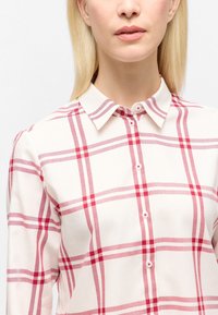 Woman wearing a white collared shirt with red plaid pattern, shown from neck to mid-torso against a plain background.