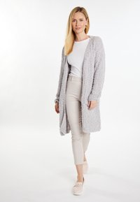 Cardigan lung, cu deschidere frontală, dintr-o textură moale de gri, purtat peste un top alb și pantaloni bej, asortat cu pantofi deschiși. Design simplu și relaxat.