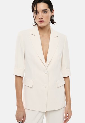 Donna con capelli corti e scuri che indossa un blazer color avorio con maniche arrotolate e pantaloni abbinati su uno sfondo chiaro uniforme.